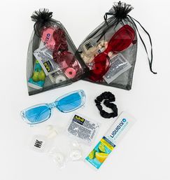 Festival survival kit flatlay with two black organza pouches holding colorful sunglasses, plus blue sunglasses, hydration powder stick, single-dose pain reliever packet, earplugs, earbuds, breath mints and a black scrunchie on white background.