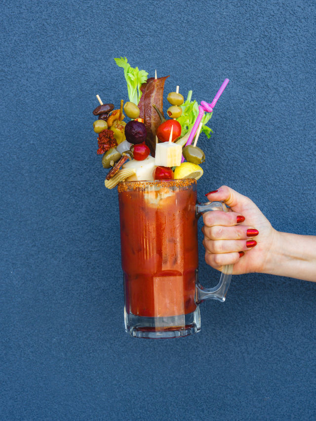 Tall Bloody Mary in a beer-style mug with a spiced rim, overflowing with skewered garnishes — celery, olives, bacon, cheese, cherry tomatoes, pickles and baby corn — pink straw and a hand with red nails holding the mug against a blue textured wall