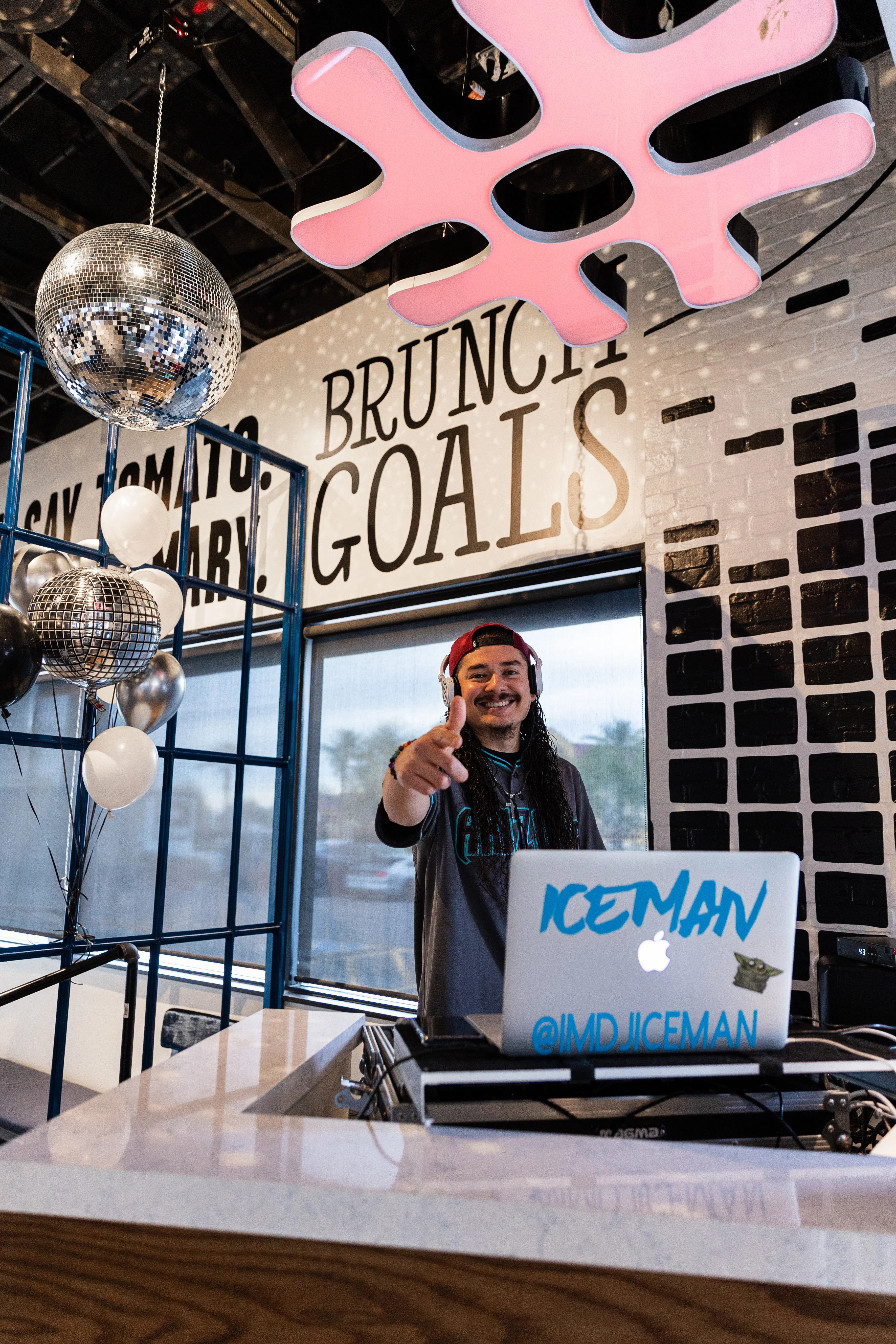 Smiling DJ with headphones points at camera from a brunch spot DJ booth, laptop, disco balls, balloons and 'BRUNCH GOALS' wall art