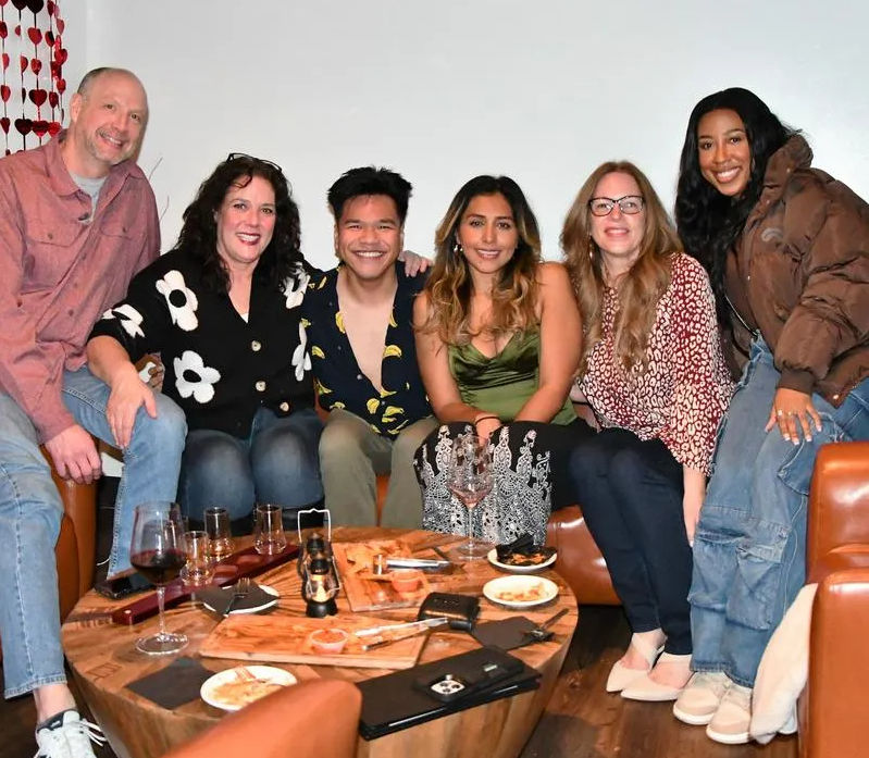 Six friends smiling on leather couches in a cozy indoor lounge, gathered around a wooden coffee table with wine glasses, small plates and a charcuterie-style spread for a casual night out.