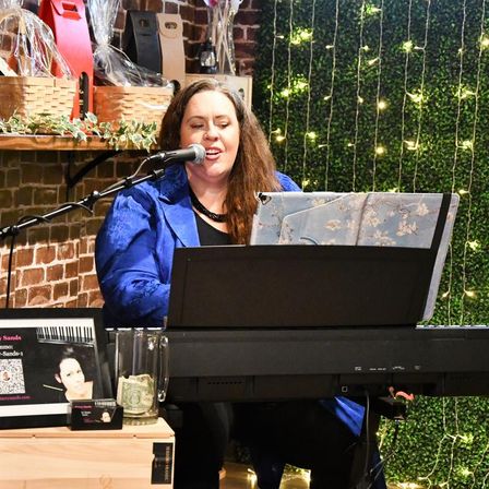 Smiling singer-pianist playing a digital keyboard and singing into a microphone at an intimate indoor venue, with exposed brick, wall greenery and warm string lights in the background.