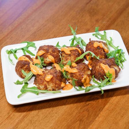 Savory, bite-sized golden croquettes on a white rectangular plate, drizzled with bright orange sauce and garnished with peppery arugula on a wooden table.