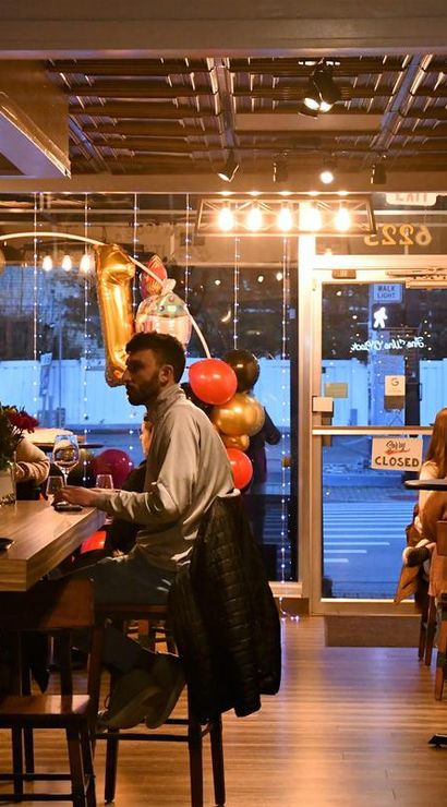 Cozy urban wine bar interior at dusk with patrons seated at a wooden bar and high tables, warm string lights, gold and red balloons including a large '1' foil balloon, glassware and bottles on shelves, and a closed storefront door visible