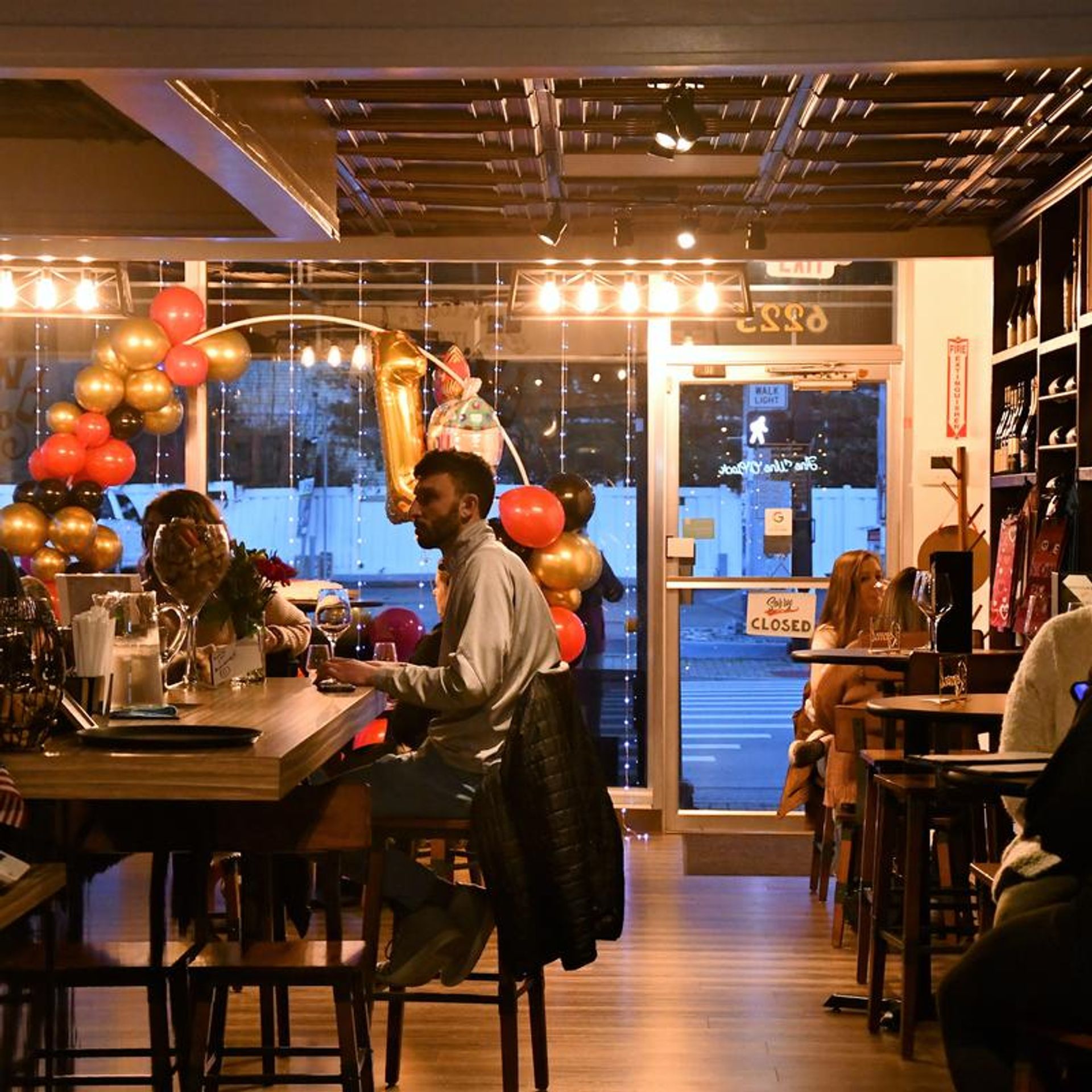 Cozy urban wine bar interior at dusk with patrons seated at a wooden bar and high tables, warm string lights, gold and red balloons including a large '1' foil balloon, glassware and bottles on shelves, and a closed storefront door visible