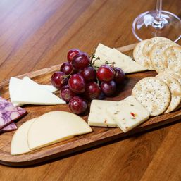 Assorted cheese board with sliced and wedge cheeses, red grapes and round crackers on a wooden serving board with a wine glass, perfect appetizer platter.