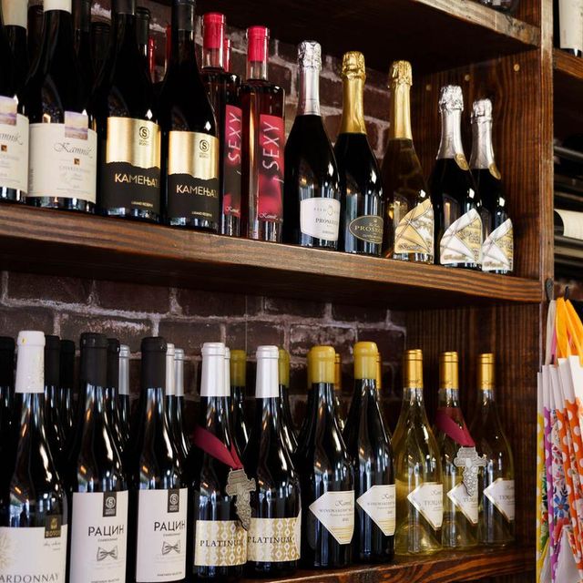 Assorted red, white and sparkling wine bottles neatly arranged on wooden shelves against an exposed brick wall in a wine shop display, showcasing foil tops and varied labels.