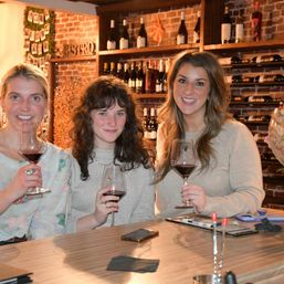 Three friends smiling and holding glasses of red wine at a cozy brick-walled wine bar with shelves of bottles behind them.
