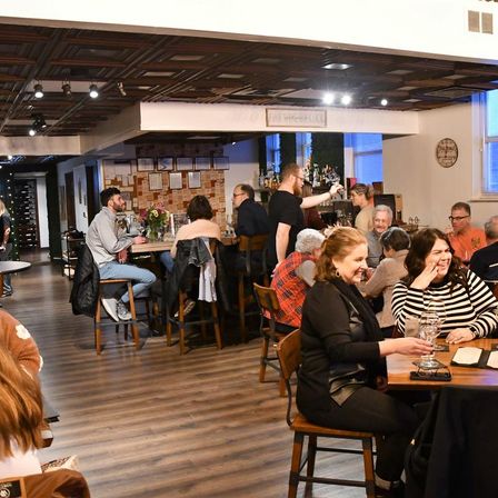 Cozy neighborhood bar and restaurant interior with wood floors and decorative ceiling; lively evening crowd chatting at high tables and bar while a bartender pours wine.