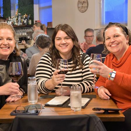 Three friends smiling and toasting with wine at a cozy neighborhood wine bar, wooden table with menus and water glasses.