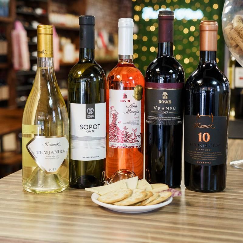 Five assorted wine bottles (two white, one rosé, two red) lined up on a wooden counter with a small plate of crackers and cheese, festive bokeh lights and wine-shop shelves blurred in the background.