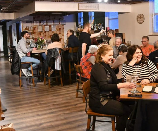 Cozy neighborhood taproom interior with wooden floors and a bar, bartender pouring drinks as groups laugh and chat at tables.