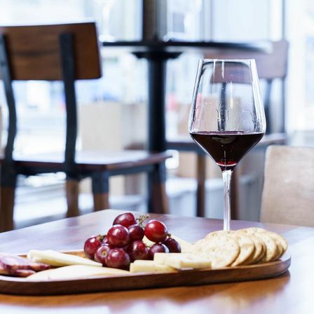 Glass of red wine with a cheese board of grapes, sliced cheeses and crackers on a wooden table in a bright café.