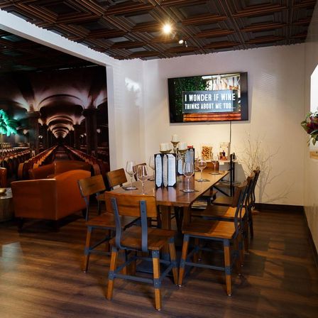 Cozy wine tasting room interior with a long rustic wooden table set with wine glasses and menus, metal‑accent chairs, hardwood floors, decorative wood ceiling, a wall TV reading 'I wonder if wine thinks about me too...' and a large wine cellar mural