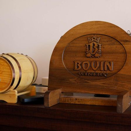 Rustic tabletop wine barrel beside an engraved wooden plaque with a winery logo on a dark wood shelf — cozy tasting-room decor.