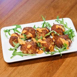 Crispy golden crab cakes drizzled with spicy orange aioli and scattered arugula on a white rectangular plate atop a wooden table, seafood appetizer