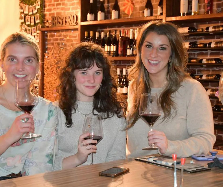 Three friends smiling and enjoying red wine at a cozy wine bar with exposed brick and shelves of bottles, smartphone on the wooden counter