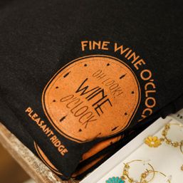 Black T-shirt folded on a boutique display featuring an orange 'Wine O'Clock' clock graphic, shown next to daisy-style gold earrings