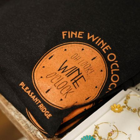 Black T-shirt folded on a boutique display featuring an orange 'Wine O'Clock' clock graphic, shown next to daisy-style gold earrings