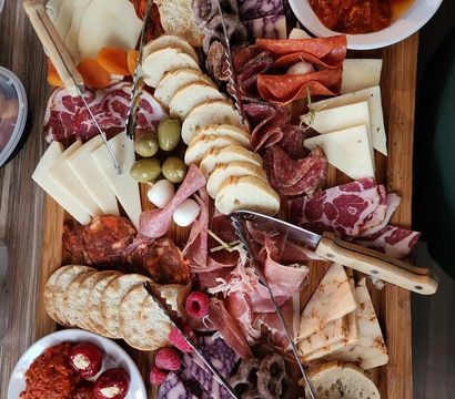 Colorful charcuterie grazing board on a wooden plank with assorted cured meats (prosciutto, salami, chorizo), sliced cheeses, crackers and baguette rounds, green olives, stuffed peppers, savory spreads, pretzels and fresh raspberries — gourmet appetizer platter for sharing.