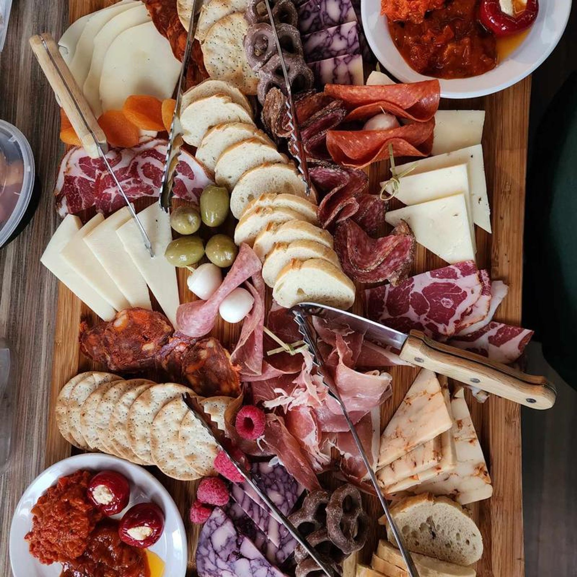 Colorful charcuterie grazing board on a wooden plank with assorted cured meats (prosciutto, salami, chorizo), sliced cheeses, crackers and baguette rounds, green olives, stuffed peppers, savory spreads, pretzels and fresh raspberries — gourmet appetizer platter for sharing.