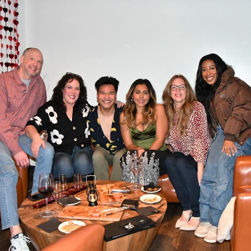 Group photo of six friends smiling on leather couches in a cozy indoor lounge, wooden coffee table with wine glasses, appetizers and a red heart garland in the background.