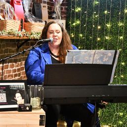 Female vocalist playing keyboard in a cozy indoor venue — microphone, sheet music, twinkle lights, brick wall and decorative greenery.