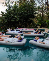 Group of women lounging on inflatable beds floating in a calm garden pool surrounded by lush tropical trees — serene spa-style wellness retreat.