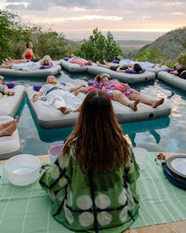 Group of people lying on inflatable mattresses in a pool at sunset for an outdoor sound-bath relaxation session, facilitator in a green robe seated at the pool edge with crystal singing bowls and a scenic hillside view.