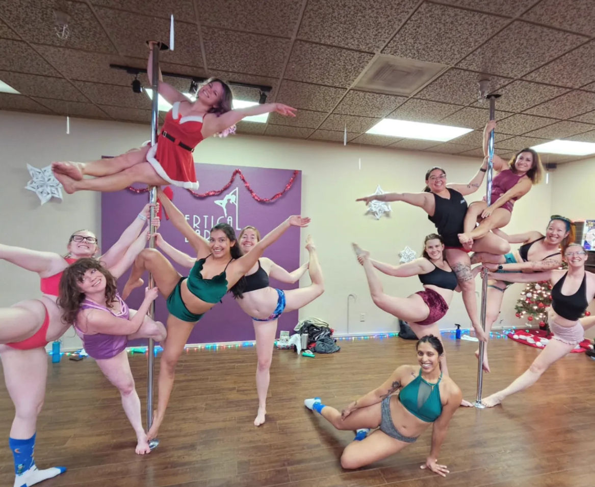 Festive pole fitness class: a smiling group of women in a dance studio performing pole and floor poses in colorful athletic and holiday outfits, posed against a purple studio wall with holiday decorations and a small Christmas tree.