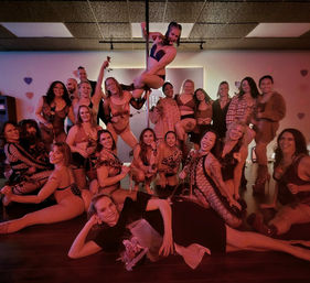 Cheerful group of women in lingerie and dancewear posing around a pole in a dimly lit dance studio with red lighting, heart decorations, roses and a bouquet — celebratory pole dance class photo.