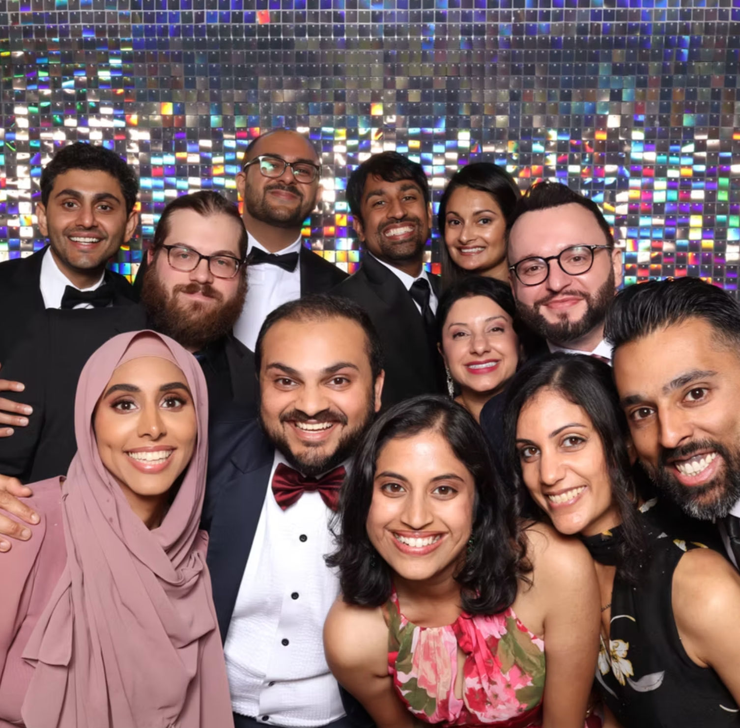 Beaming group of friends in black-tie attire posing tightly in a sparkly sequin photo‑booth backdrop at a formal party.