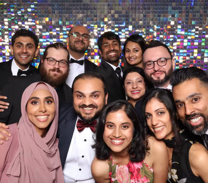 Beaming group of friends in black-tie attire posing tightly in a sparkly sequin photo‑booth backdrop at a formal party.