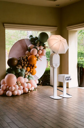 Playful pastel balloon installation in peach, sage, orange and gold around a circular backdrop beside a white photo booth with umbrella light on a covered tile patio entrance.