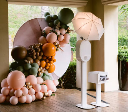 Playful pastel balloon installation in peach, sage, orange and gold around a circular backdrop beside a white photo booth with umbrella light on a covered tile patio entrance.