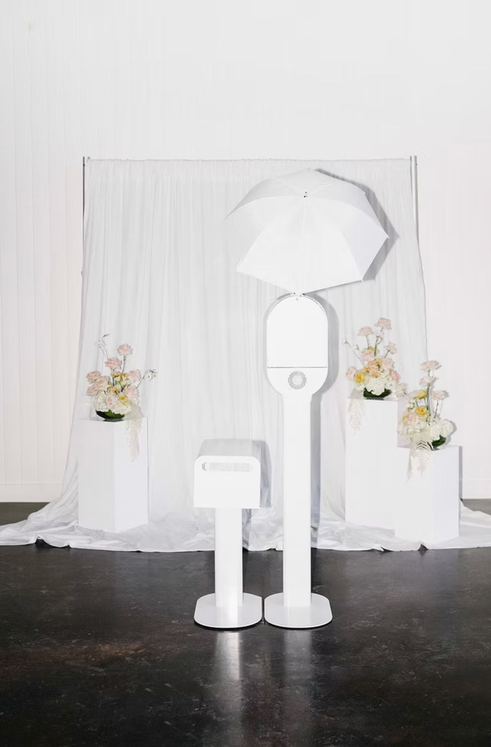 Minimal white photo booth with umbrella light and small printer in front of a white draped backdrop, flanked by pedestal floral arrangements — modern wedding or event studio setup