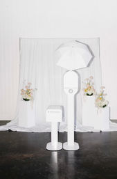 Minimal white photo booth with umbrella light and small printer in front of a white draped backdrop, flanked by pedestal floral arrangements — modern wedding or event studio setup