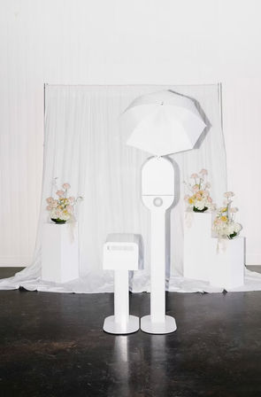 Minimal white photo booth with umbrella light and small printer in front of a white draped backdrop, flanked by pedestal floral arrangements — modern wedding or event studio setup