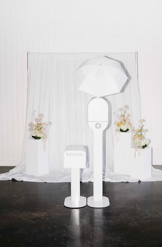 Minimal white photo booth with umbrella light and small printer in front of a white draped backdrop, flanked by pedestal floral arrangements — modern wedding or event studio setup