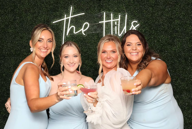 Four smiling women — bride in white and three bridesmaids in light-blue dresses — cheering with cocktails at a wedding reception photo booth in front of a green hedge wall and neon script sign.