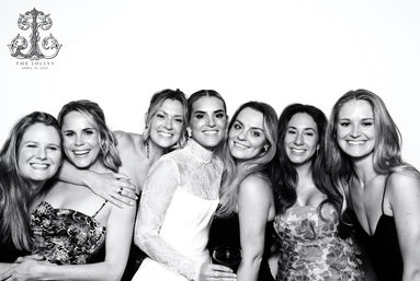 Black-and-white wedding photo booth portrait of a smiling bride in a long-sleeve lace gown surrounded by six laughing bridesmaids and friends, a joyful close-knit group posing against a white backdrop.