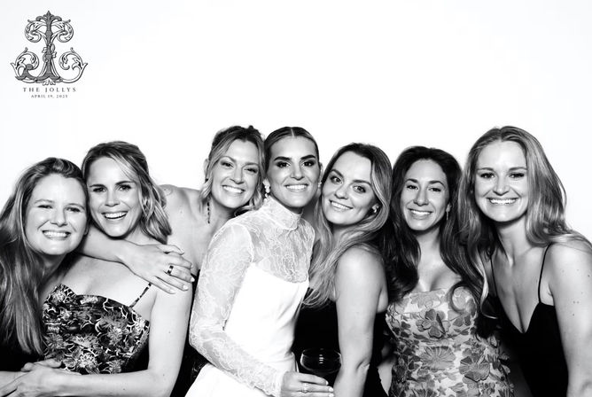 Black-and-white wedding photo booth portrait of a smiling bride in a long-sleeve lace gown surrounded by six laughing bridesmaids and friends, a joyful close-knit group posing against a white backdrop.