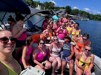Smiling group of friends in colorful swimsuits on a pontoon boat enjoying a sunny summer lake day, holding drinks with lake houses and docks visible in the background.