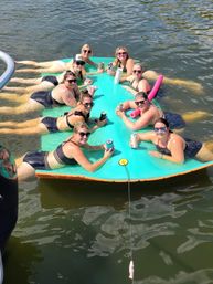 Smiling group of friends in swimsuits lounging on a bright teal floating mat in a lake, holding canned drinks and tumblers on a sunny summer day.