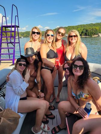 Eight women in swimsuits and sunglasses enjoying a sunny summer pontoon boat party on a forest-lined lake, holding drinks with a purple ladder and blue sky in the background.