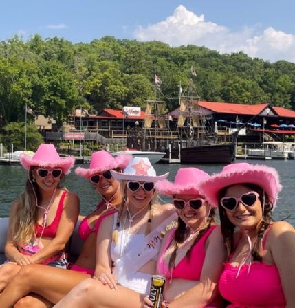 Five women in pink cowboy hats and heart-shaped sunglasses on a lake boat, celebrating with one wearing a white sash, pink swimsuits, waterfront docks and a tall pirate-style ship in the background under a sunny blue sky.