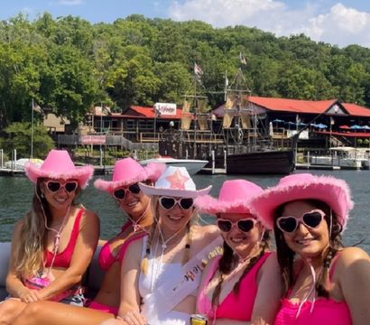 Five women in pink cowboy hats and heart-shaped sunglasses on a lake boat, celebrating with one wearing a white sash, pink swimsuits, waterfront docks and a tall pirate-style ship in the background under a sunny blue sky.