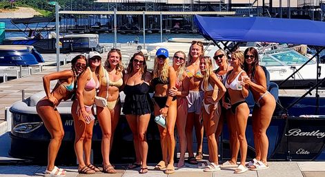 Group of friends in bikinis posing on a sunny lake marina dock in front of a pontoon boat, enjoying a summer boating day