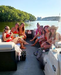 Seven friends in swimsuits enjoying a sunny summer pontoon-boat outing on a calm lake with tree-lined shore and distant waterfront buildings.
