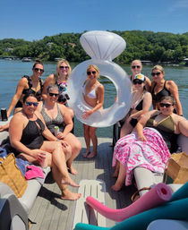 Smiling group of women in swimsuits on a pontoon boat at a sunny lake, holding a giant inflatable diamond-ring float with tree-lined shoreline and other boats in the background.