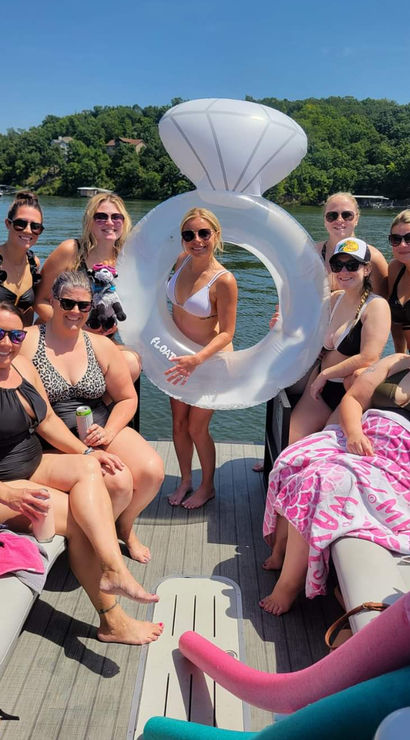Smiling group of women in swimsuits on a pontoon boat at a sunny lake, holding a giant inflatable diamond-ring float with tree-lined shoreline and other boats in the background.